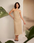 Woman wearing a beige halter neck dress standing against a neutral background with green leaves.