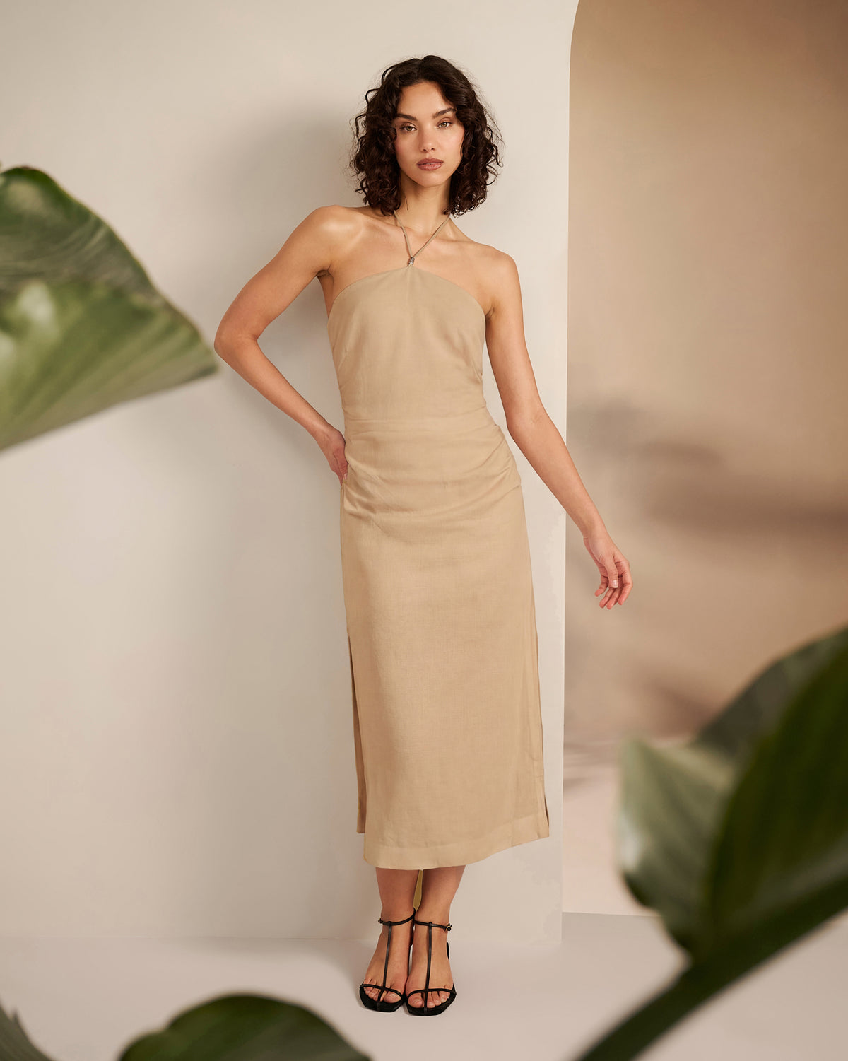 Woman wearing a beige halter neck dress standing against a neutral background with green leaves.